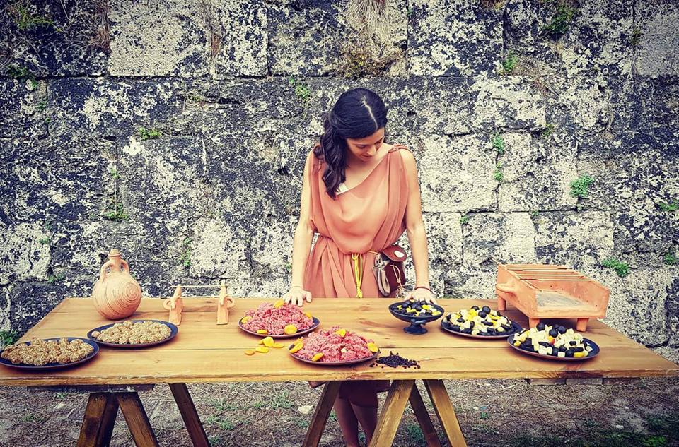 Historical Gastronomy Preparation | Archaeopolis Archive Woman in historical dress arranging reconstructed dishes on a wooden table beside an ancient stone wall