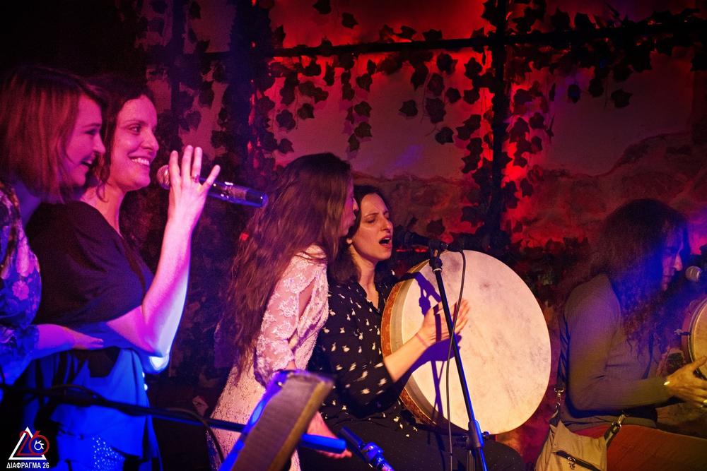 Women singing and playing a large frame drum during a red-lit stage performance