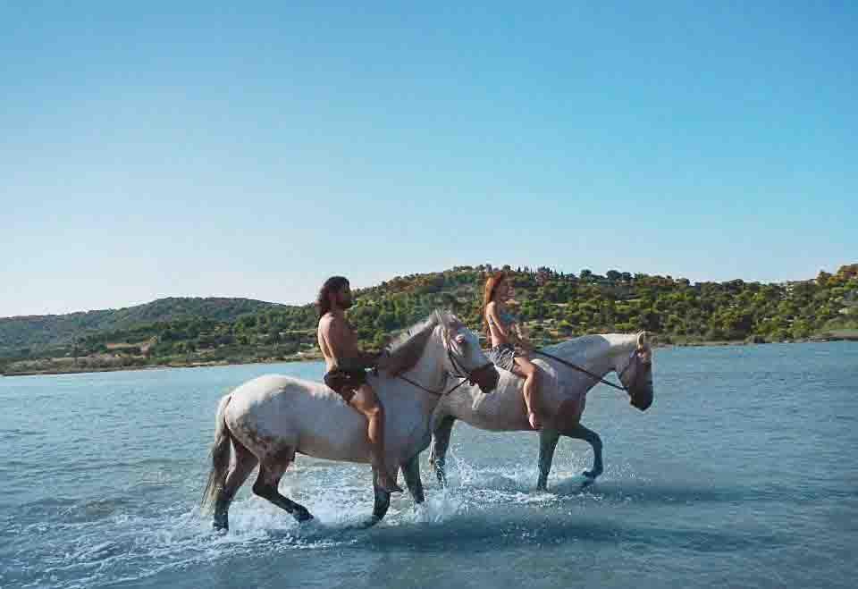 Two women riding horses together through clear shallow coastal water