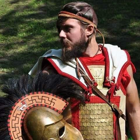 Hoplite interpreter outdoors holding a decorated bronze helmet and round shield