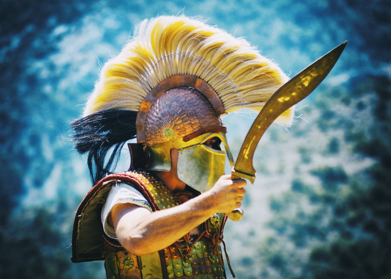 Armoured Greek warrior wearing a high-crested bronze helmet and raising a curved sword against a blue landscape