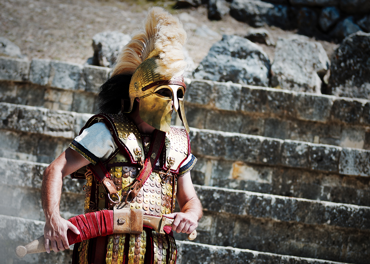 Armoured hoplite wearing a crested bronze helmet while drawing a sword before ancient stone seating