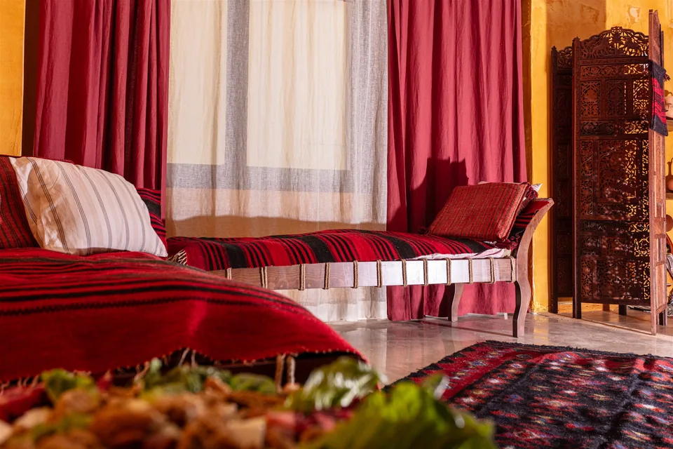 Wooden reclining couches dressed with red and black woven covers and cushions, beside draped curtains and a carved folding screen