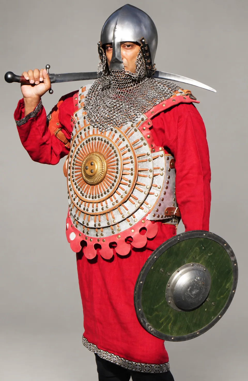 Modern mirror-armour interpretation with concentric chest plates, mail, red tunic, sword and round shield against a neutral background