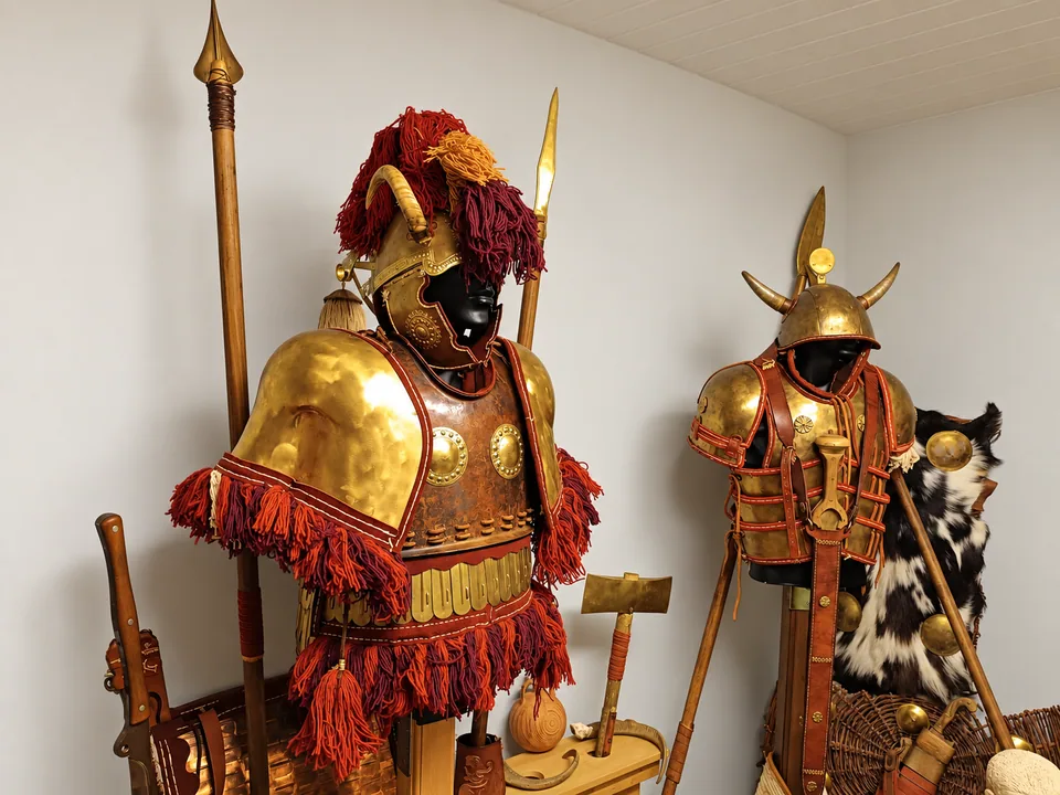 Angled view of the Mycenaean equipment display with two armour ensembles, horned helmets, spears, axes and a hide-faced shield