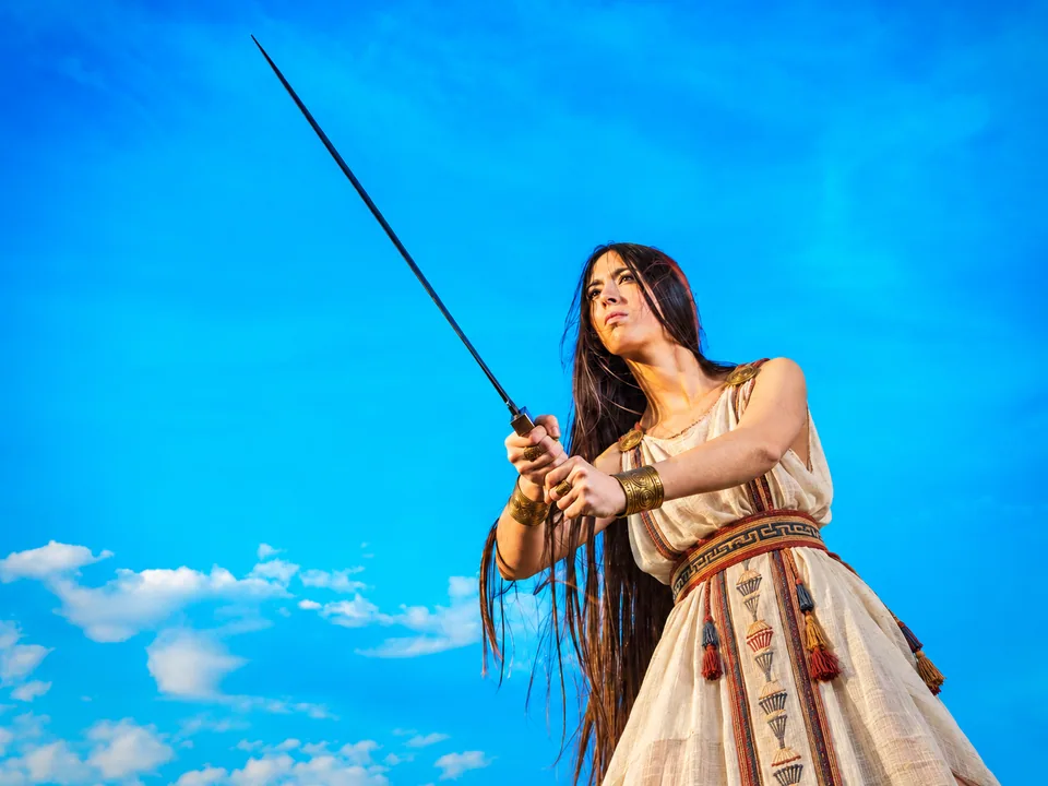 Woman holding a raised sword with both hands, wearing a cream dress with patterned borders, a woven belt, circular shoulder fastenings and wrist cuffs against a blue sky