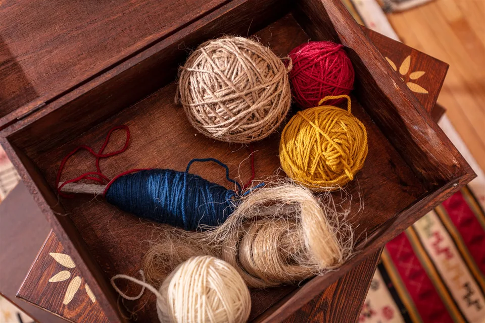Wooden tray holding red, yellow and cream yarn, natural fibres and blue thread wound around a wooden tool