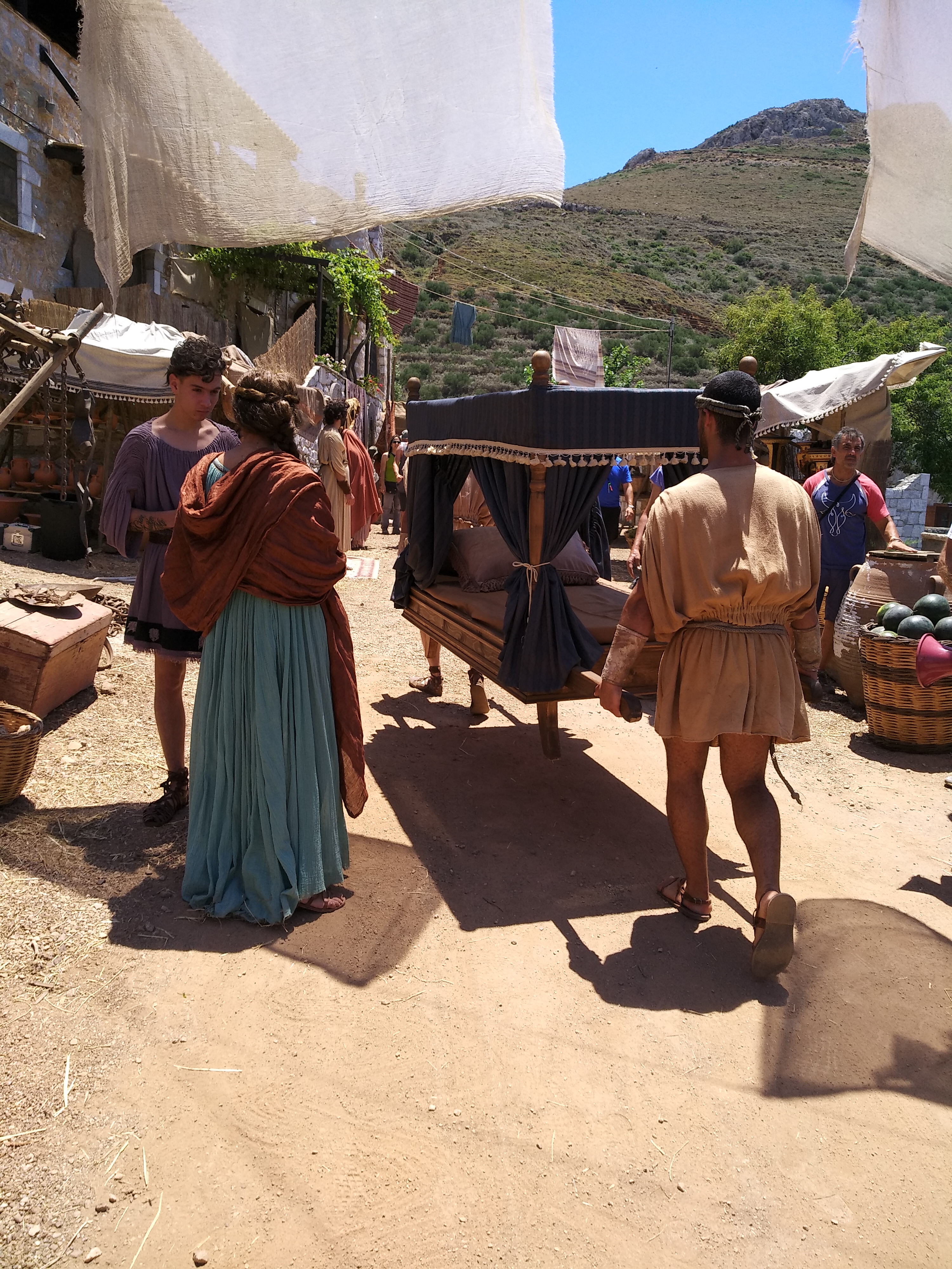 Assassin’s Creed Odyssey promotional production · agora environment behind the scenes Behind-the-scenes view of the Assassin’s Creed Odyssey agora set with performers, market stalls, pottery, textiles and a carried bed