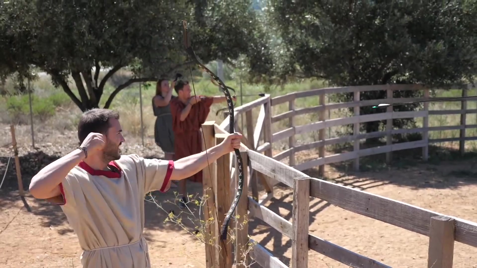 Archery practice with a shield target | Archaeopolis Archery Games Costumed archer releasing toward a shield target in a fenced arena