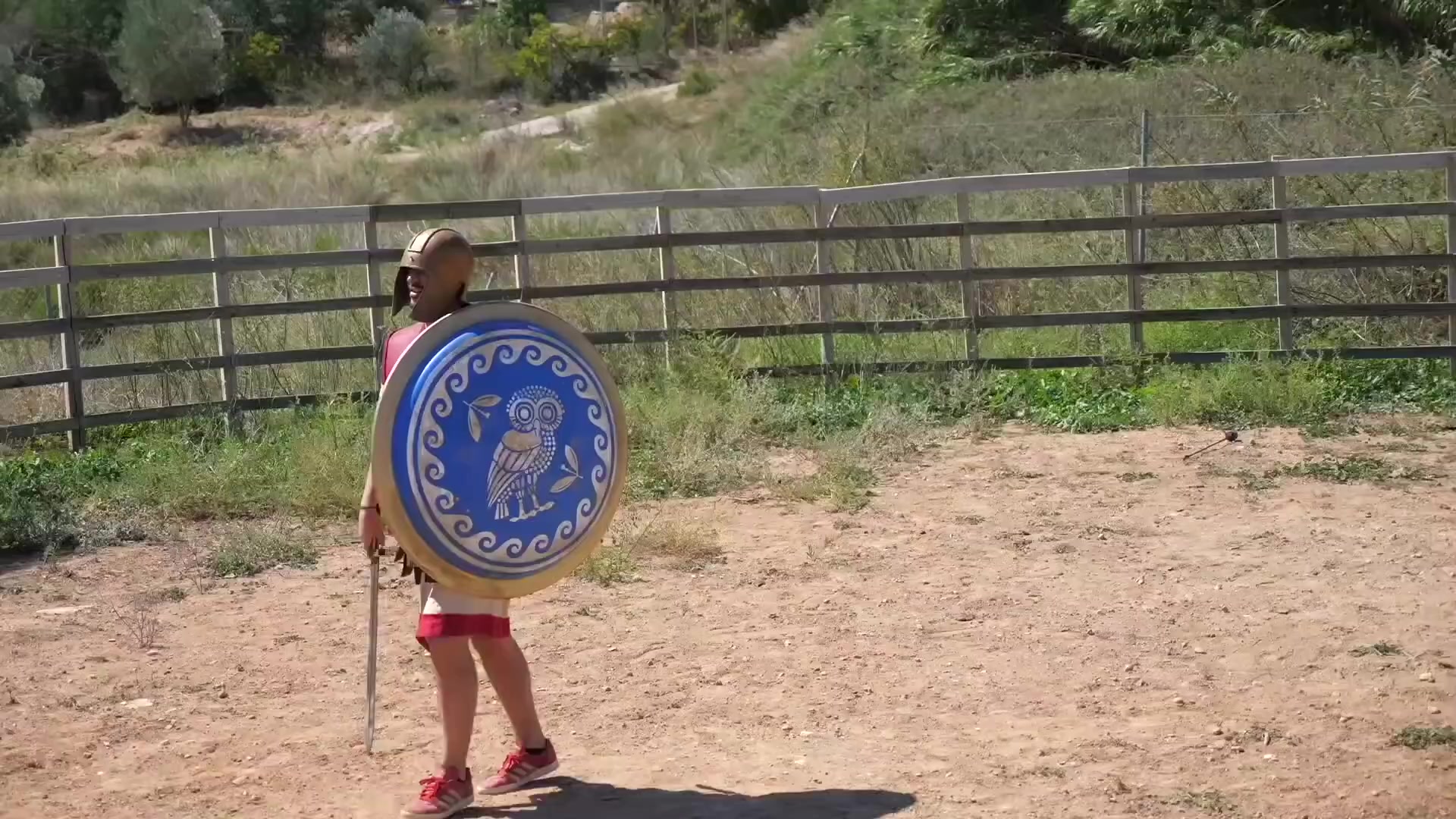 Shield-bearing participant in the games arena | Archaeopolis Archery Games Costumed participant carrying the blue owl shield across the arena