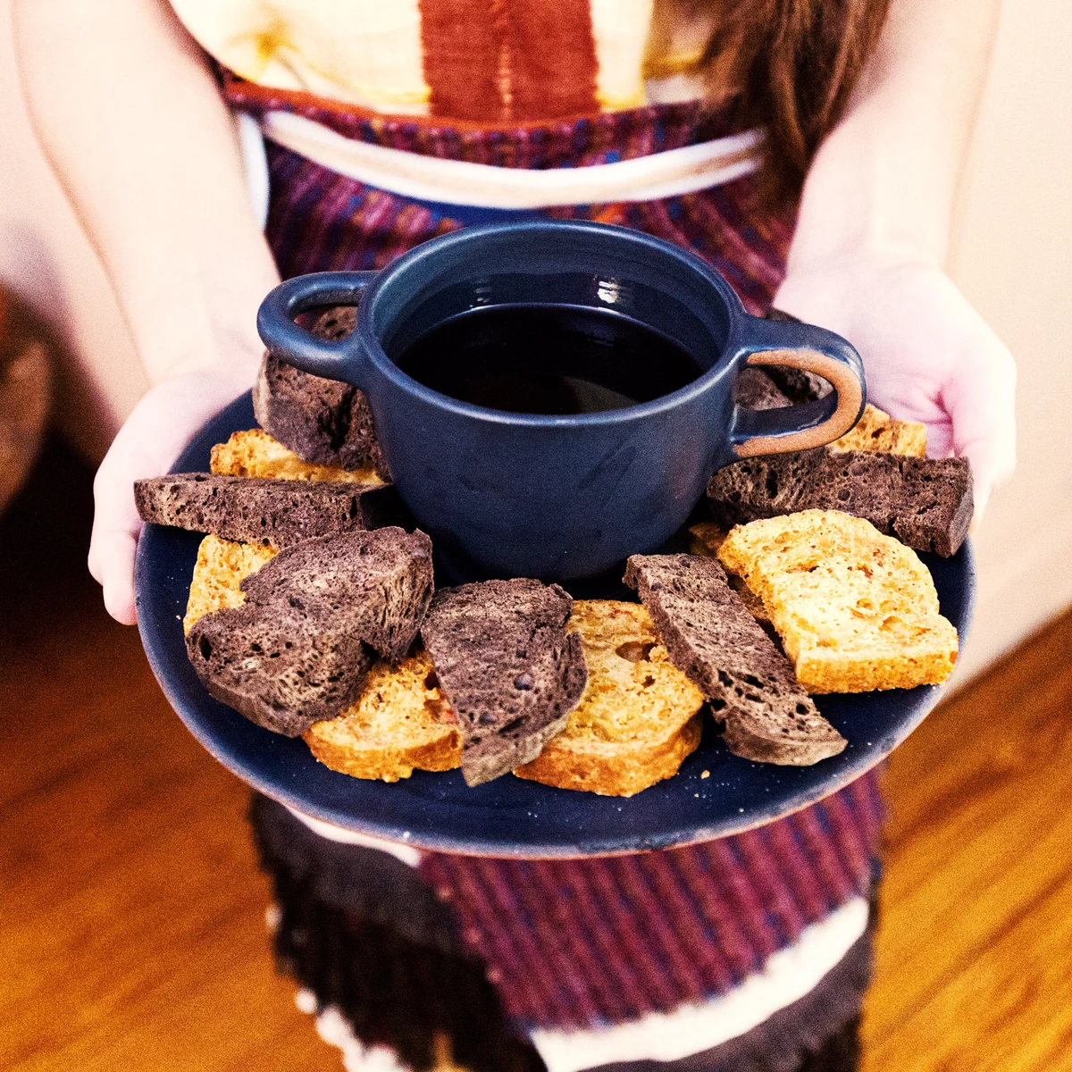 A platter of sliced bread surrounding a dark two-handled cup, held out for serving.