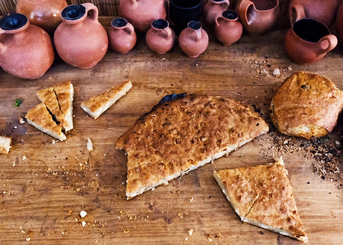 Bread and sliced baked preparations on a wooden table beneath terracotta jugs and cups.