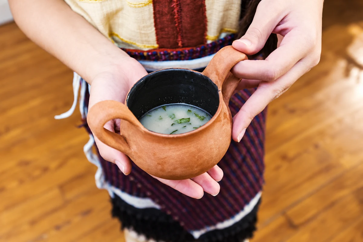 A two-handled terracotta cup containing a pale drink with green herbs, held in two hands.