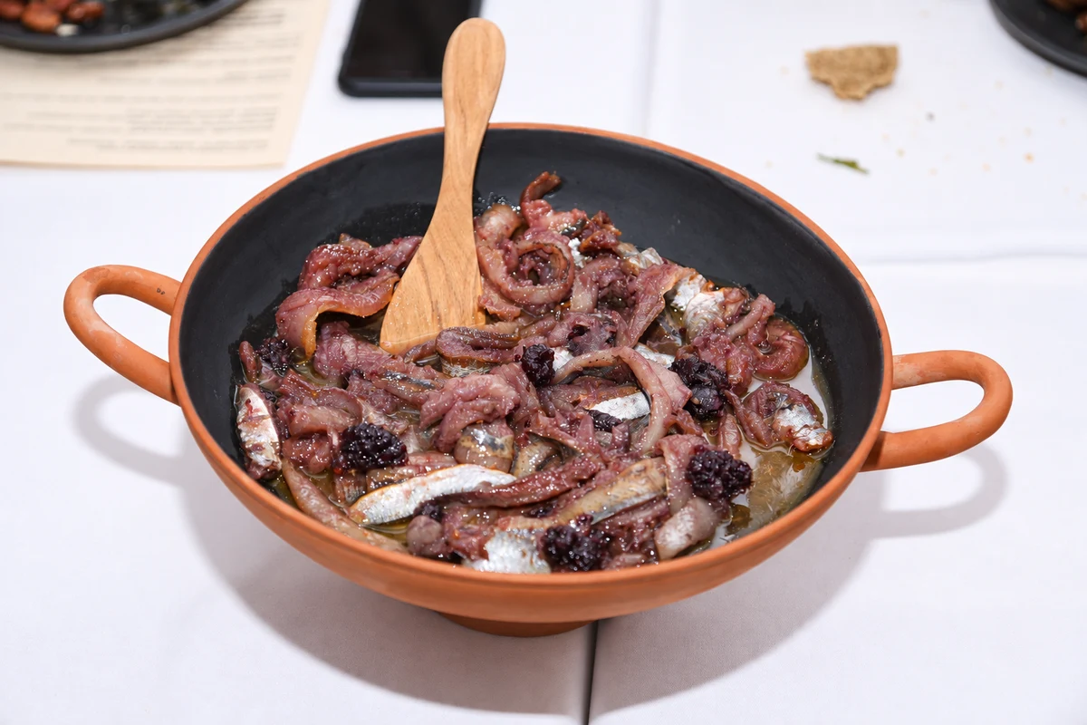 Small fish with sliced onions and dark fruit in a terracotta bowl with a wooden spoon.