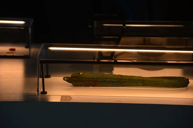Close-up of a long green-patinated metal blade inside a horizontal museum case.