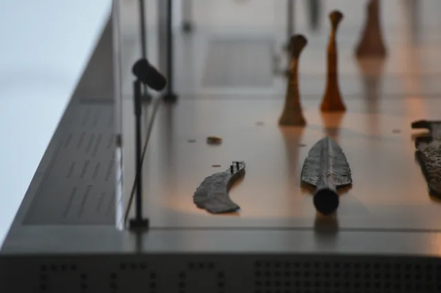 Ancient metal spearhead and smaller metal fragment displayed with elongated objects behind them.