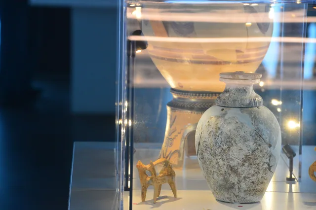 Pale ceramic vessel and small animal figurine displayed before a larger painted pot.