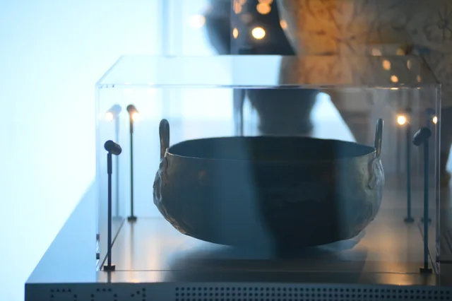 Large dark metal vessel with two upright handles displayed under museum lighting.