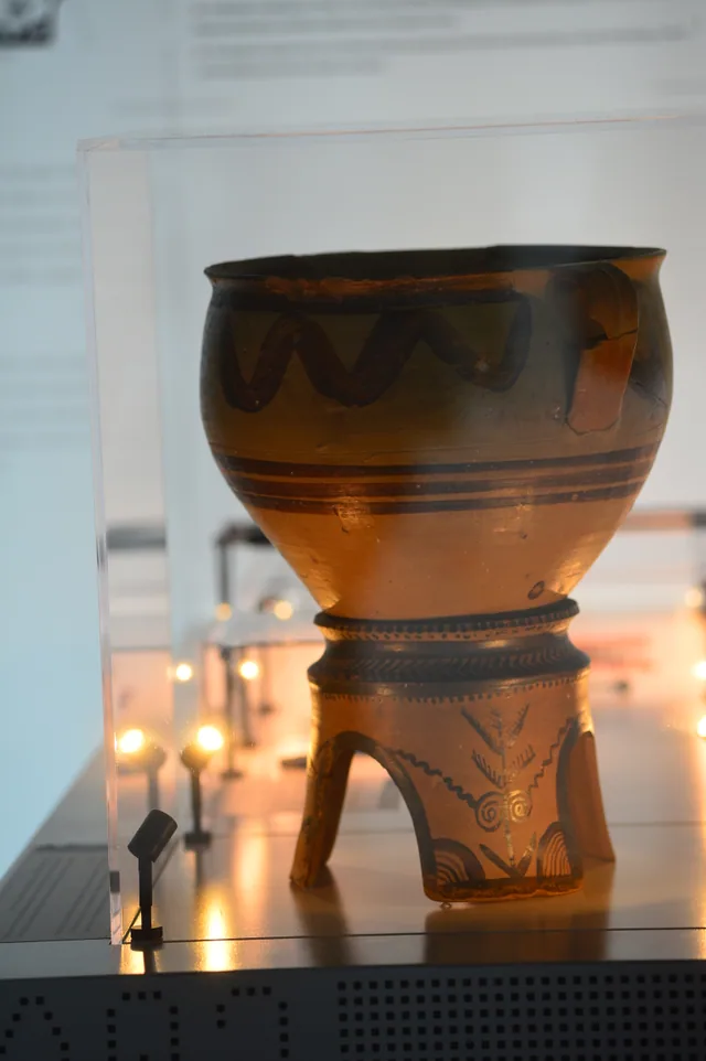 Large painted ceramic vessel with wave patterns on a decorated tripod stand, photographed from an angle.