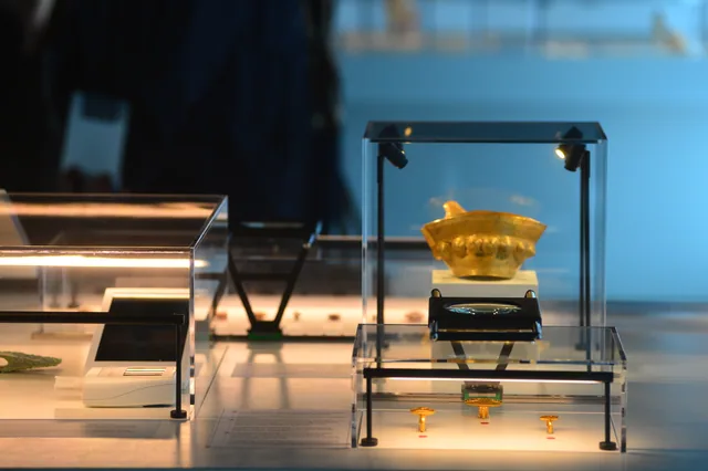 Small gold vessel with three gold objects and a magnifying lens inside a display case.