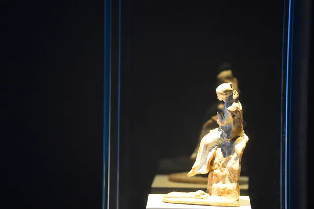 Pale seated terracotta human figurine shown in side view against a dark museum background.