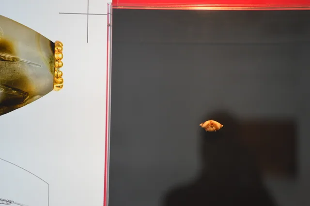 Tiny gold ornament displayed against a black background beside an exhibition graphic.
