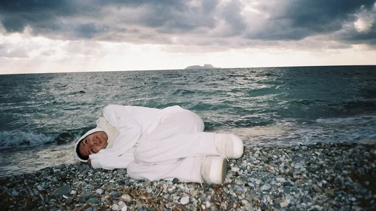 Official LAB12 exhibition image showing a white-clad figure lying beside the sea