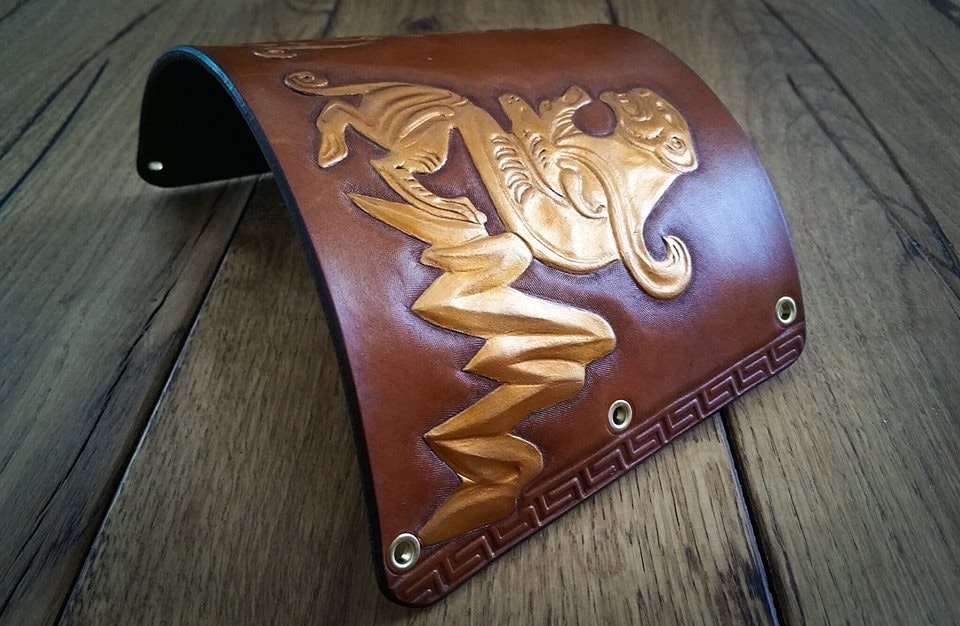 Decorated leather archery armguard with a gold-toned motif, photographed on a wooden surface