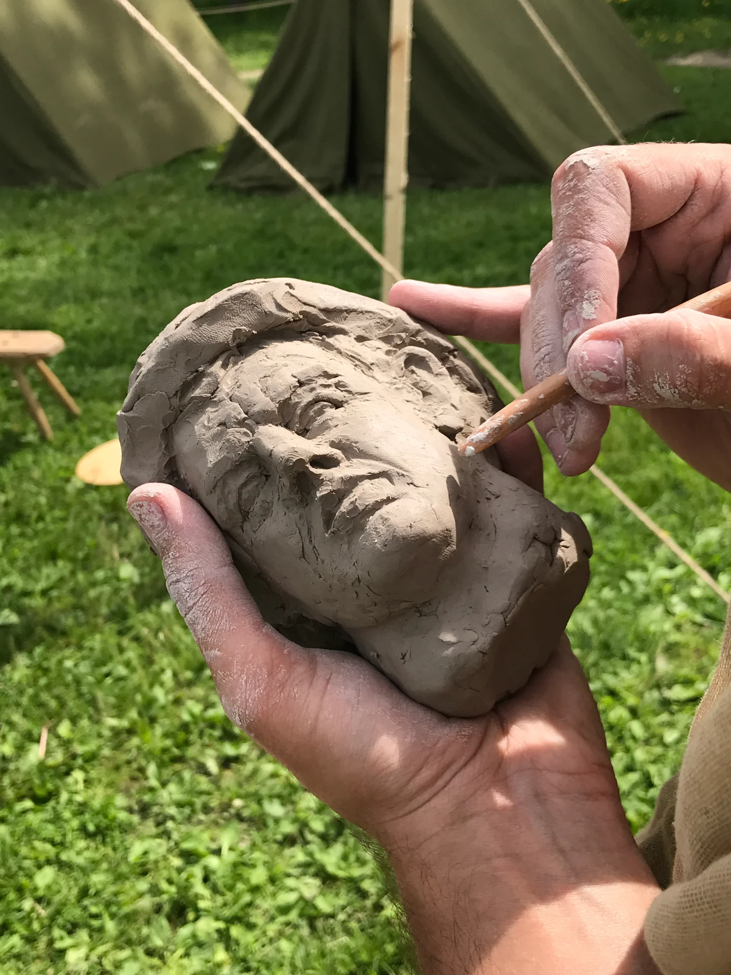 Hands shaping a small clay portrait with a wooden modelling tool outdoors