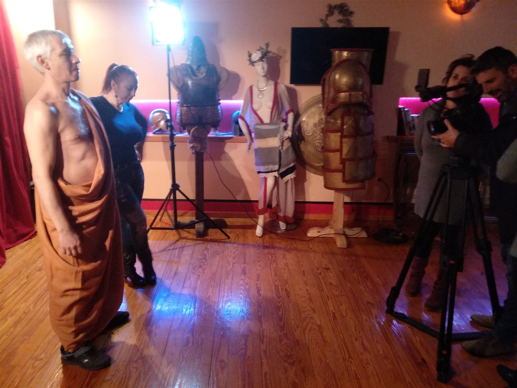 Male model in a draped garment beside a COSMOTE camera crew, reconstructed dress displays and Archaeopolis armour