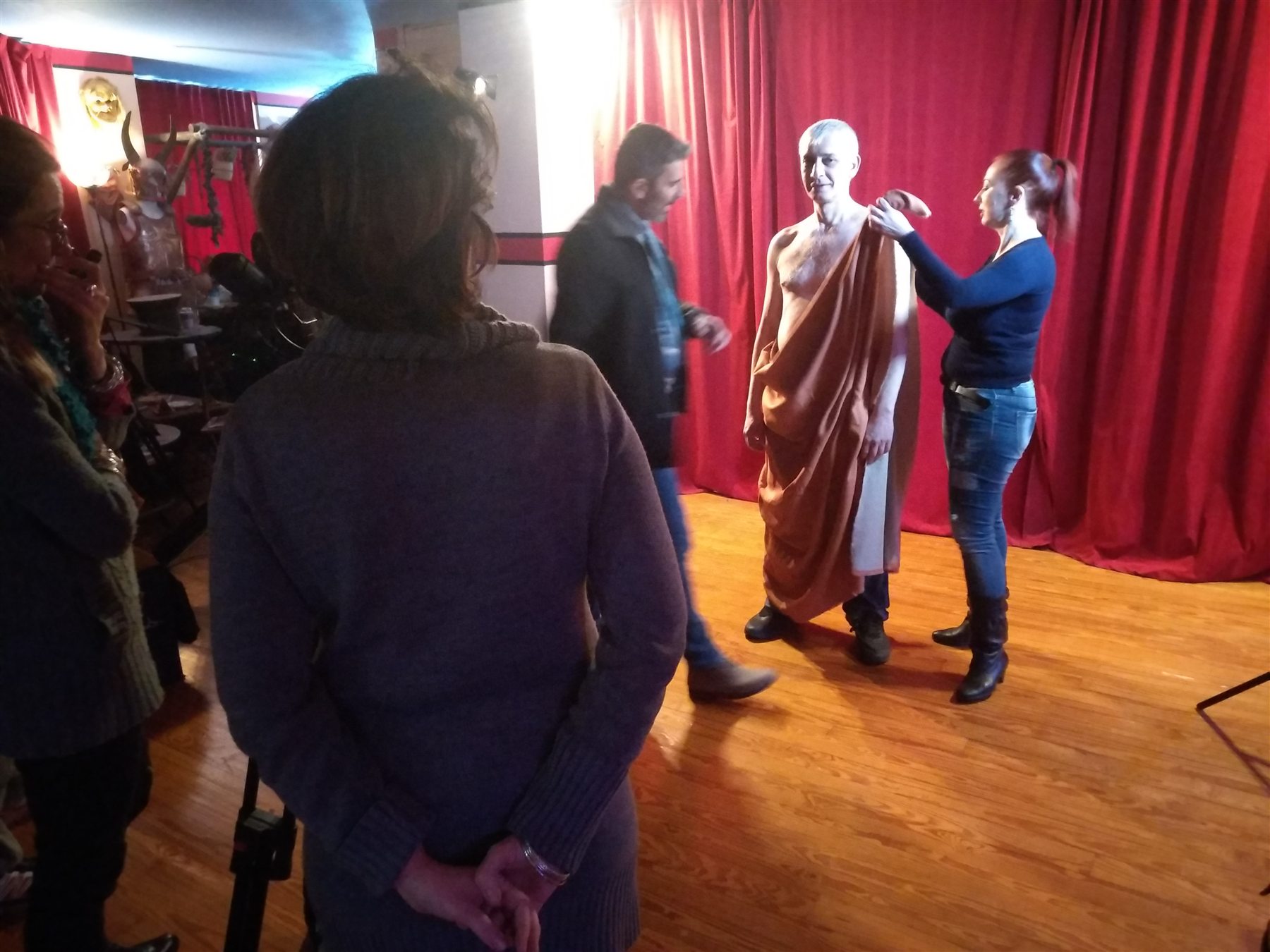 COSMOTE Ancient Greek Dress filming — 004 Final adjustment of a male model's draped garment in front of red studio curtains