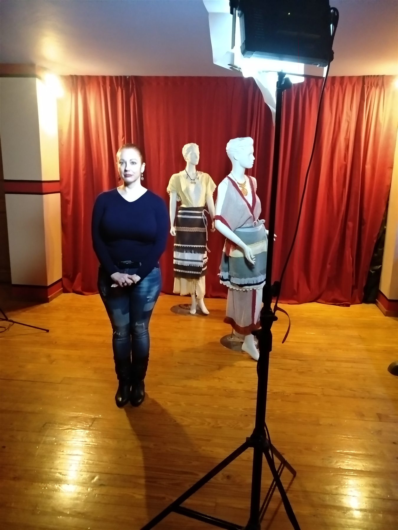 COSMOTE Ancient Greek Dress filming — 008 Presenter standing before layered reconstructed garments displayed on mannequins