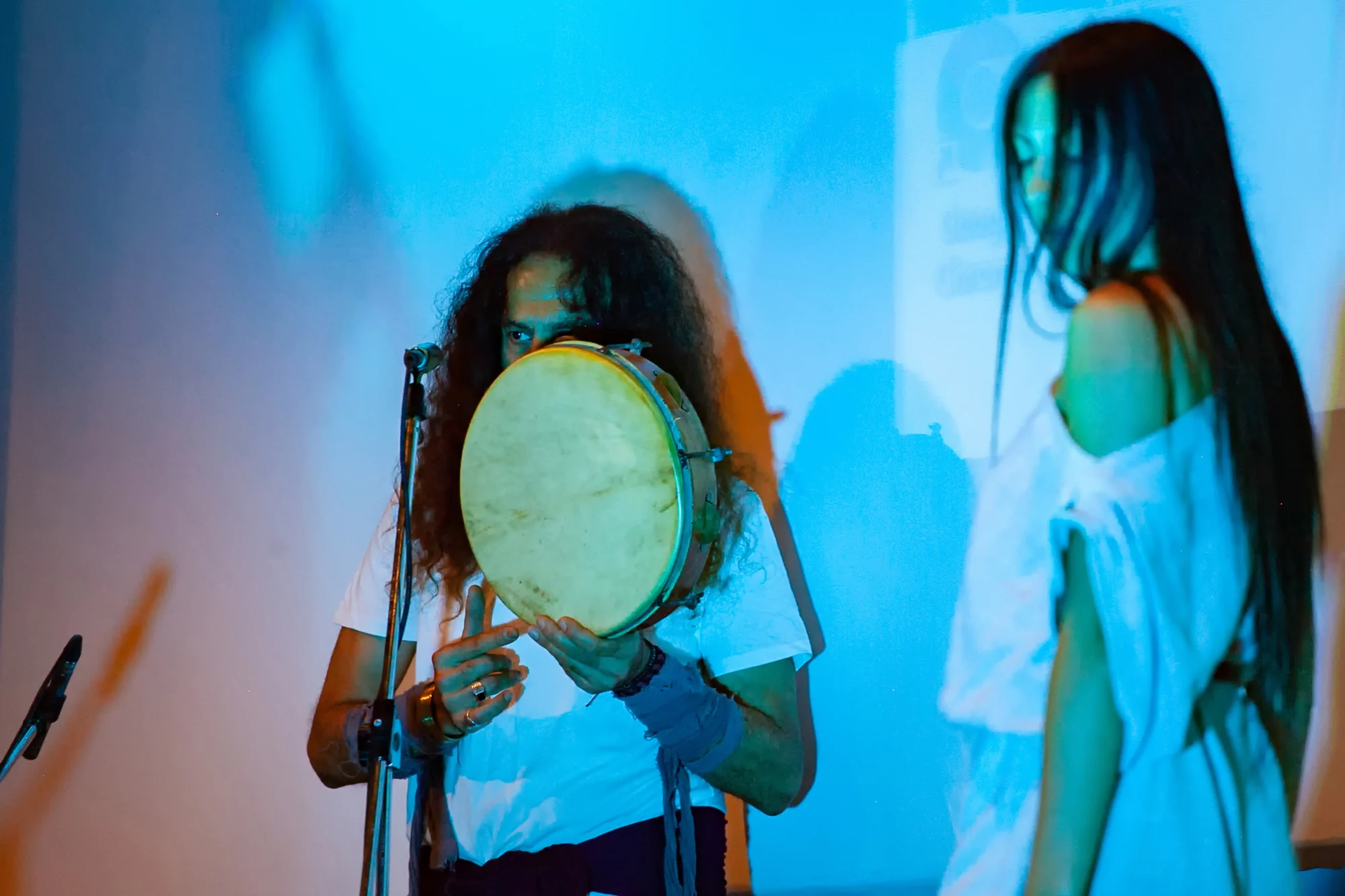 A musician holds a frame drum beside a dancer under blue stage lighting.
