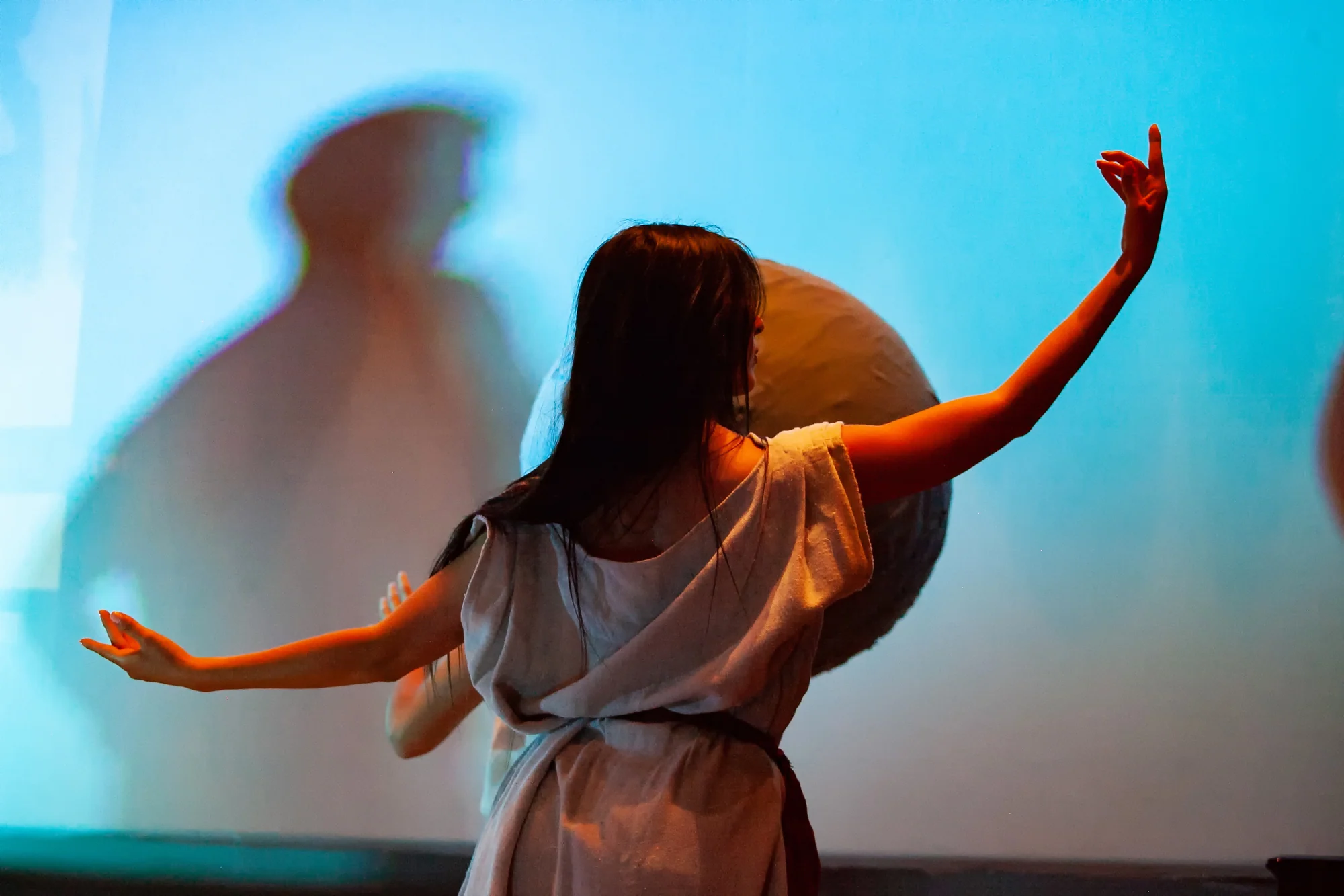 A dancer extends both arms in front of the globe prop and a large shadow on the blue-lit backdrop.