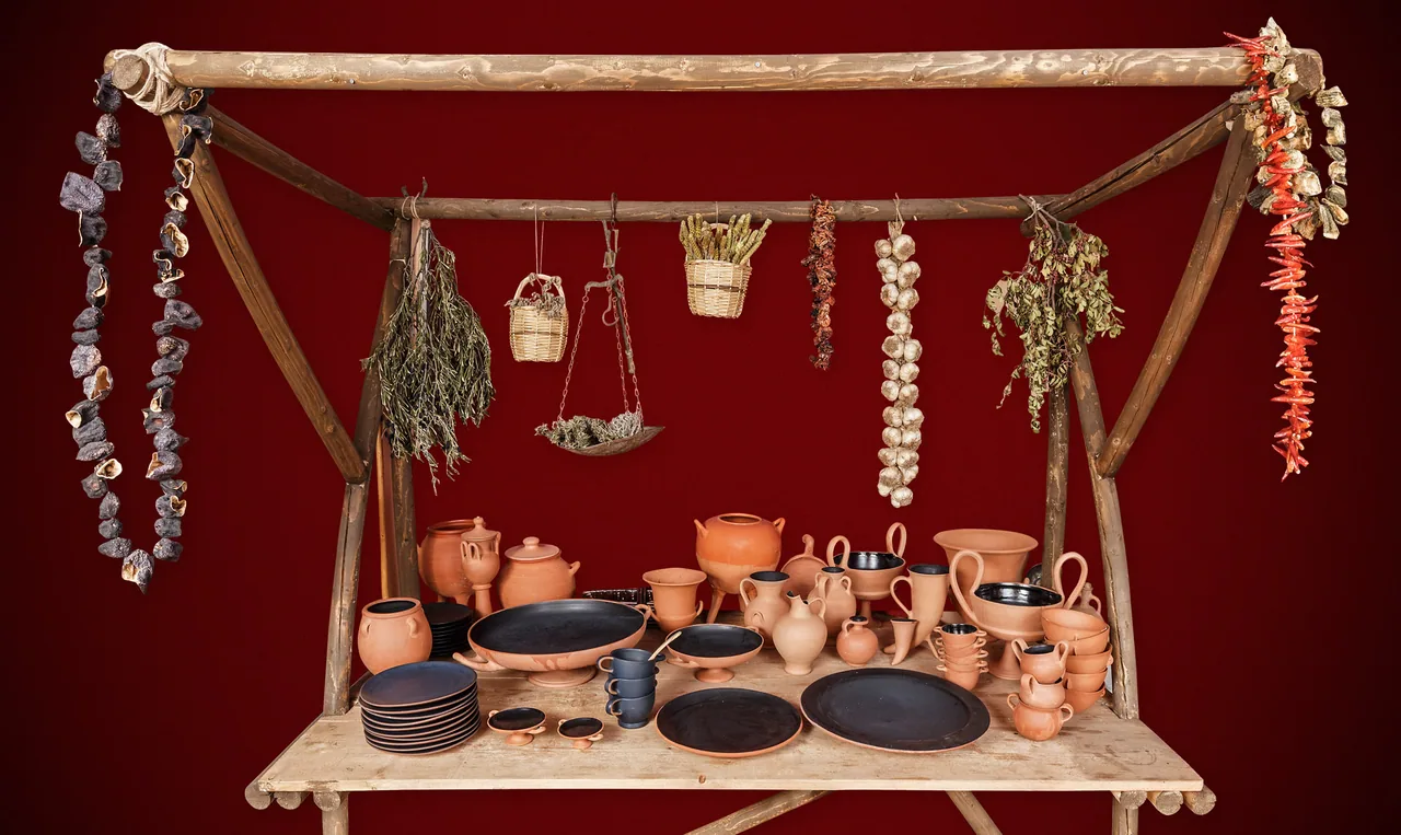 Ceramic vessels, plates and cups on a wooden market stall with hanging herbs and baskets against a red background
