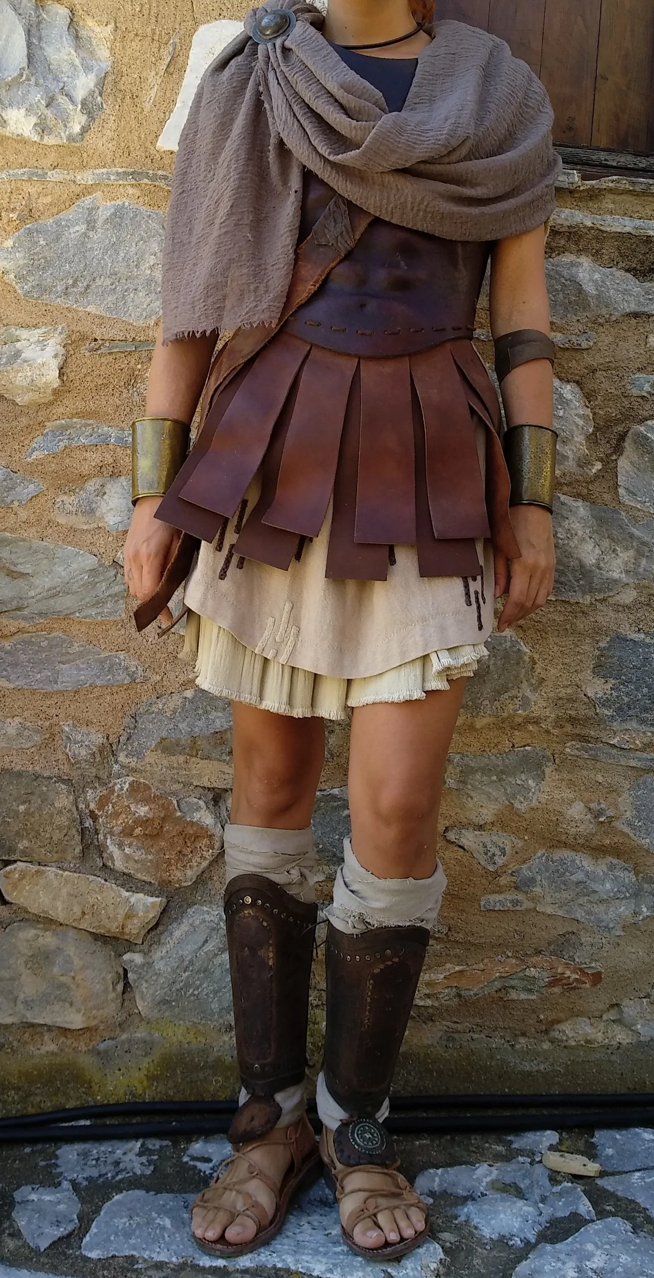 Costume detail showing a brown cuirass with layered hanging strips, a grey cloak fastened at the shoulder, pale garments, bronze-coloured wrist cuffs, shin guards and sandals against a stone wall