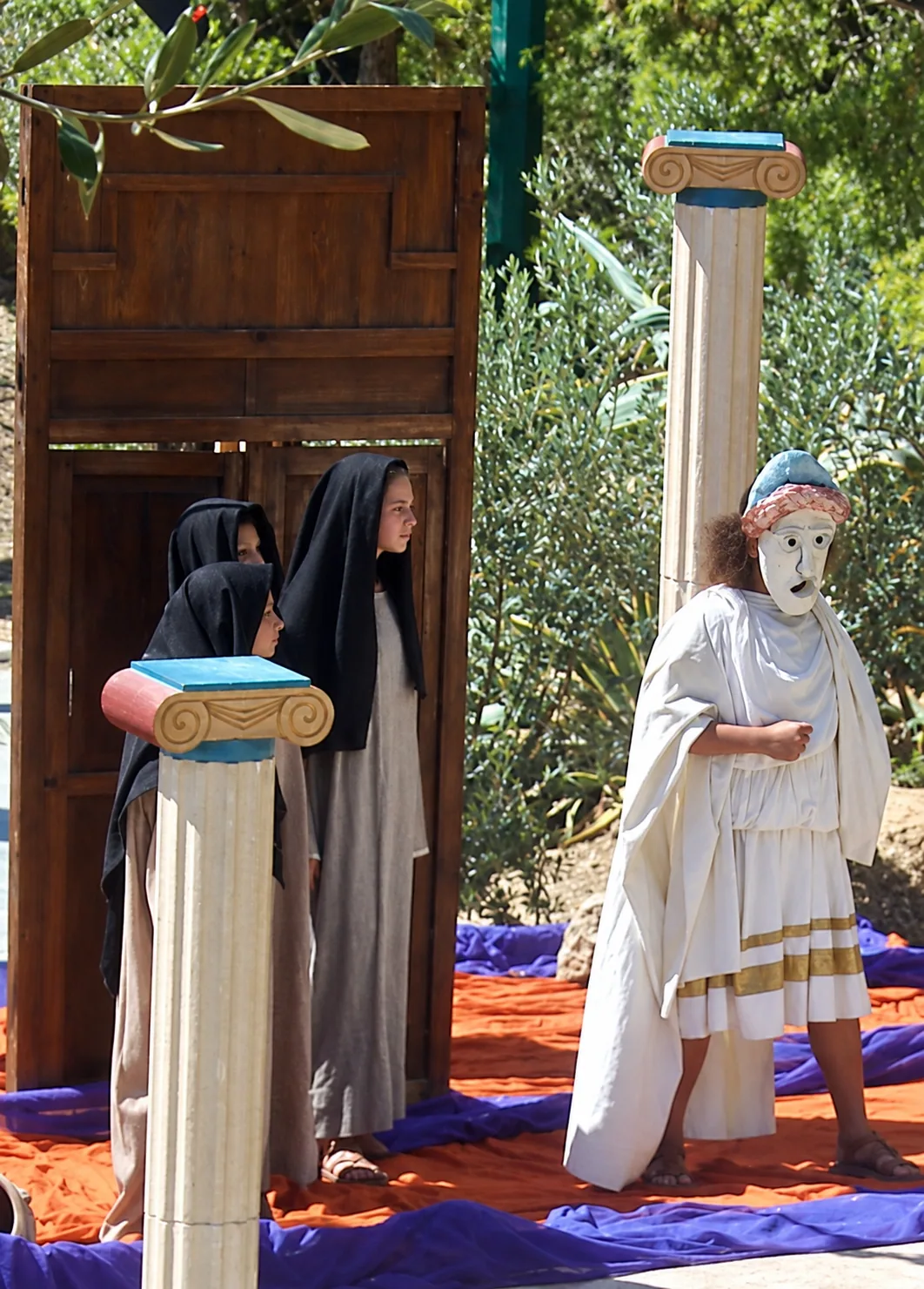 Masked performer in white draped costume beside a cloaked chorus and Ionic stage columns outdoors
