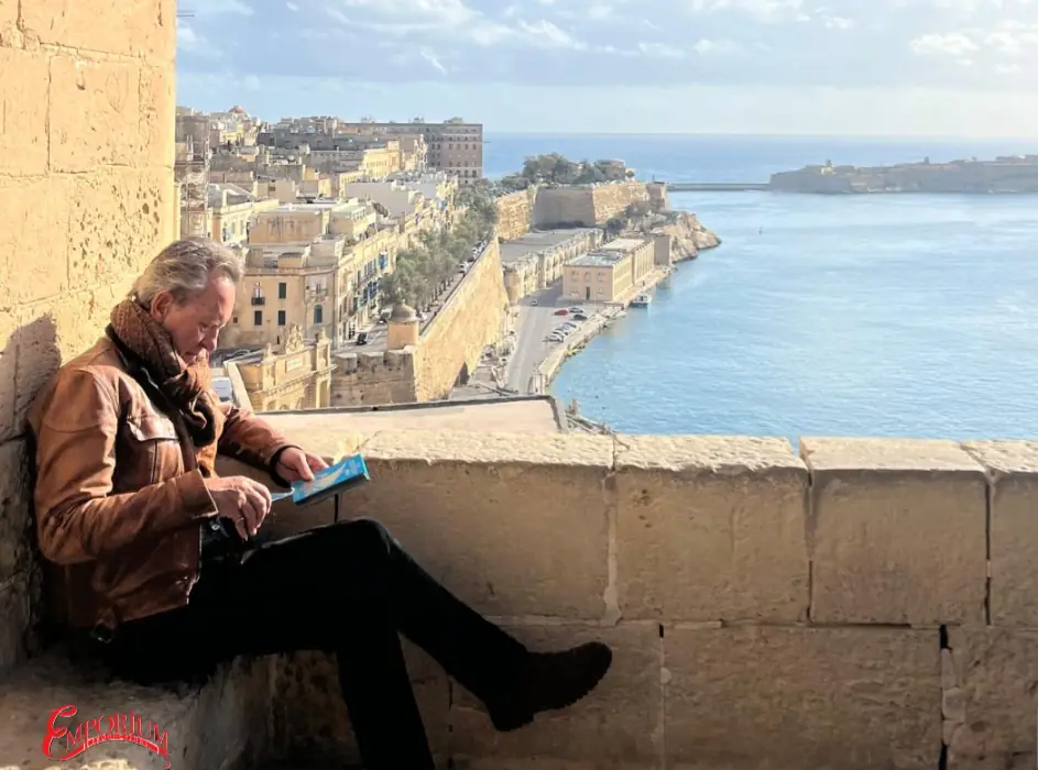 Official publicity still of Richard E. Grant reading beside a Mediterranean harbour